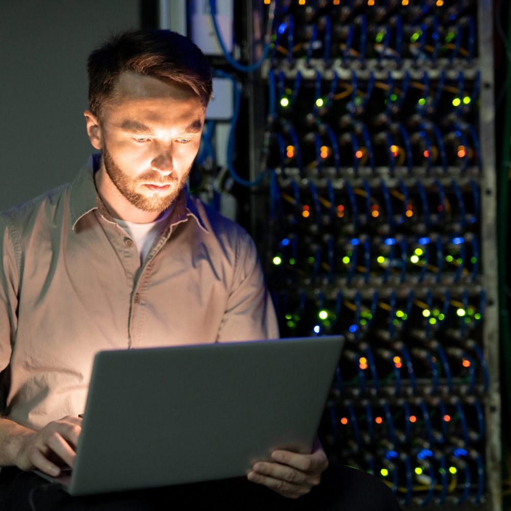 Concentrated young bearded server manager in shirt working with laptop in dark room of data center